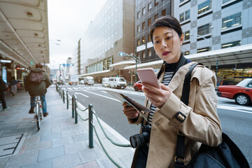 korean female traveler standing at bus shelter is checking information on phone against manual....