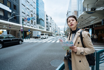 chinese traveler looking afar with map and smartphone is searching for guideboard on main road. woman tourist holding paper guide and phone and looking into distance wants to know correct direction.