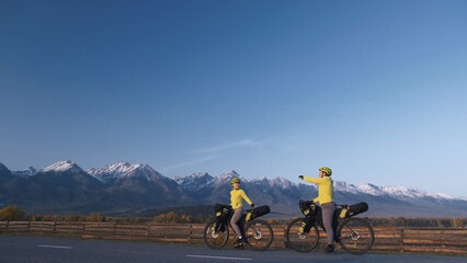The man and woman travel on mixed terrain cycle touring with bikepacking. The couple journey with bicycle bags. Sport hike bikepacking, bike, sportswear in green black colors. Mountain snow capped.