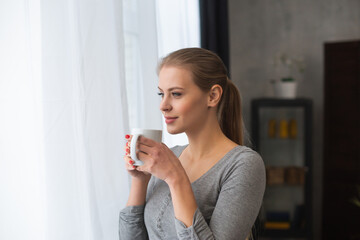 Young teenage girl is standing in front of the window and looking into it. Blond woman at home.