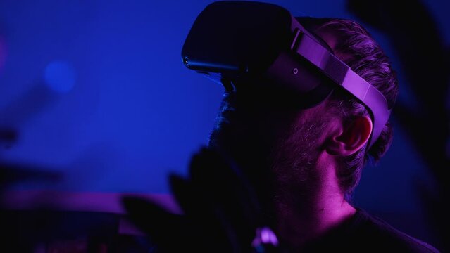 Close-up Of A Bearded Man Wearing VR Glasses In A Dark Room With Blue Light. There Is A Room Flower In The Front Captivity. VR Helmet For Entertainment, Learning And Gaming