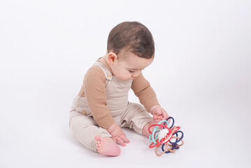 Adorable baby boy wearing beige overalls over white background holding and playing with his baby teething. 