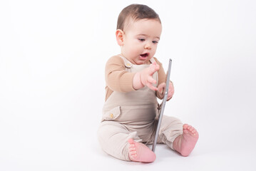 Adorable baby boy wearing beige overalls sitting on white background holding a tablet pad and watching streaming videos or cartoons. 