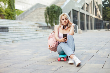 Fototapeta premium Young smiling cool girl sitting on skateboard and using phone in the city