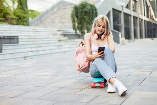 Young smiling cool girl sitting on skateboard and  using phone in the city