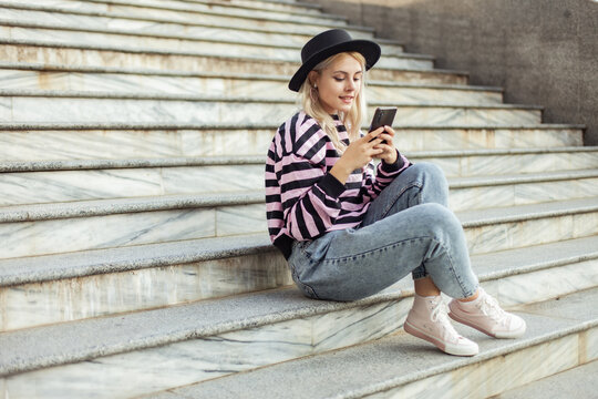 Young charming hipster girl in hat using phone while sitting on the stairs outdoors