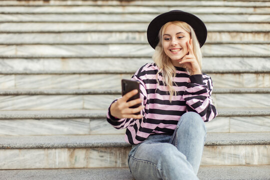 Young charming hipster girl in a hat makes a selfie on the phone while sitting on the stairs outdoors