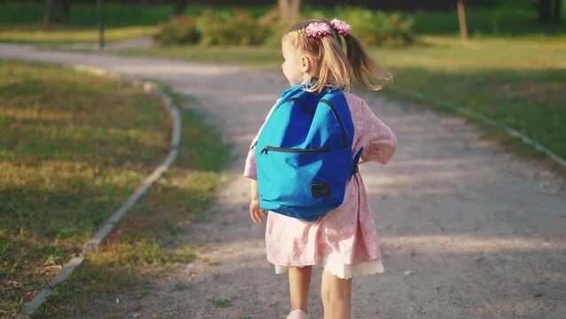 back to school. a little girl with a backpack runs through the green park to school along the path. education kid sun dream concept. a small child with a backpack back view runs the park to school