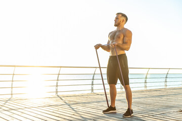 Obraz premium Young athletic man with a naked torso works out with fitness rubber bands in the early morning on the beach. Muscle training. Healthy lifestyle