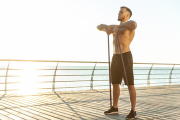 Young athletic man with a naked torso works out with fitness rubber bands in the early morning on the beach. Muscle training. Healthy lifestyle