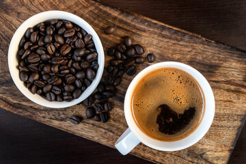 Close-up of hot black coffee in white coffee cup and roasted coffee beans in white bowl on wooden background. Top view 