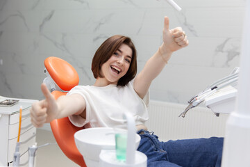 Beautiful smiling emotional woman showing thumb up while sitting in chair at dental clinic