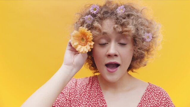 Playful curly girl is playing with a sunflower looking at camera. Portrait in yellow background