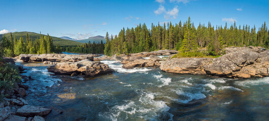 Naklejka na ściany i meble Panoramic northern landscape with rapids and cascades of wild river Kamajokk, boulders and spruce tree forest and hills in Kvikkjokk village in Swedish Lapland. Summer sunny day, blue sky white clouds