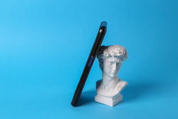 Antique bust of david with smartphone on blue background. Minimal still life.