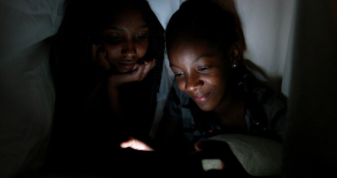 Two sisters under blanket at night watching video on smartphone device