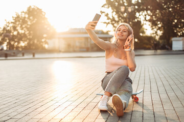Young smiling cool girl sitting on skateboard and makes selfie on phone in the city