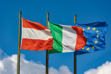Italian, Austrian, and European Union flags with flagpole, blowing togetherness in the wind on a clear blue sky with clouds and copy space.