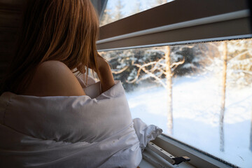 Unrecognizable girl in bed and leaning against the window looking out at the snowy exterior. 
