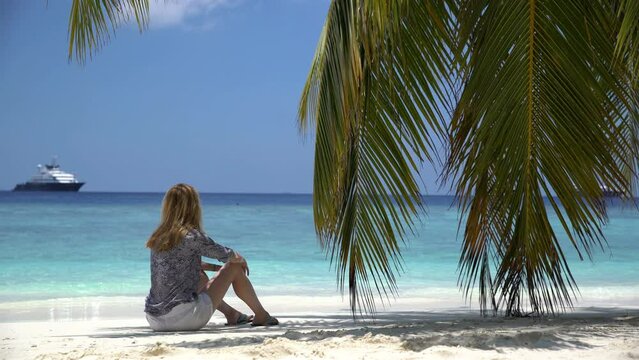 Young woman sitting by the ocean under a palm tree.