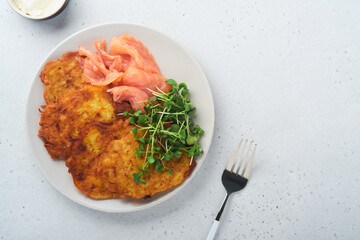 Potato pancakes. Fried homemade potato pancakes or latkes with cream, green onions, microgreens, red salmon and sauce in rustic plate on white table background. Rustic style. Healthy food. Top view.