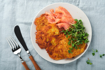 Potato pancakes. Fried homemade potato pancakes or latkes with cream, green onions, microgreens, red salmon and sauce in rustic plate on blue linen tablecloth. Rustic style. Healthy food. Top view.