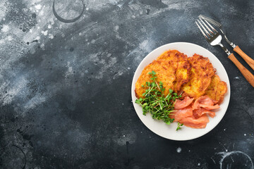 Potato pancakes. Fried homemade potato pancakes or latkes with cream, green onions, microgreens, red salmon and sauce in rustic plate on old black table background. Rustic style. Top view.