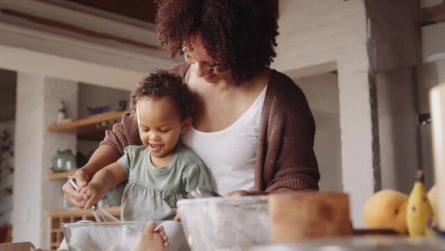 Biracial mother whisking egg with her daughter while baking in a modern-styled kitchen.