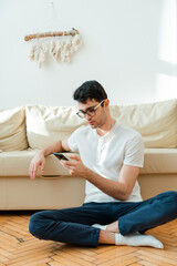 Portrait of an attractive, smiling man in casual clothes sitting on the floor near the sofa in the living room and watching the news using a mobile phone.