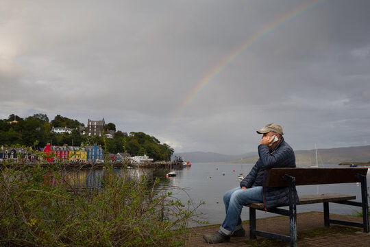 A man sits on a bench making a phone call on a mobile phone.He wears a baseball hat.In background is sea and coloured buildings of Tobermory in Scotland