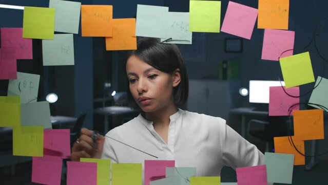Pensive businesswoman work on new strategy on glassboard with notes in dark office
