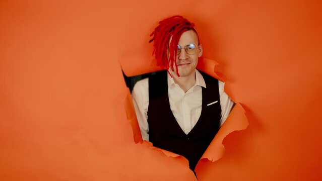 Smiling man with red dreadlocks in hole in wall. Stylish male with bright dreadlocks looking at camera through hole in orange wall in studio