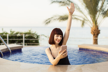 Joyful Caucasian girl in black swimsuit, with smartphone in hands, relaxing in resort sea pool on sunny day, smiling and raising hand up. People, vacations and gadget concept