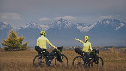 The man and woman travel on mixed terrain cycle touring with bikepacking. The two people journey with bicycle bags. Sport bikepacking, bike, sportswear in green black colors. Mountain snow capped.