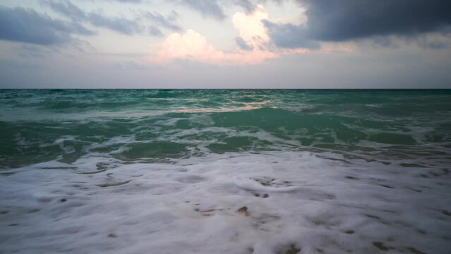 low shot of blue green waves rolling on sandy beach with white foam and the red tinged sunset clouds reflected in the sea shot at vijay nagar beach havelock andaman nicobar island India