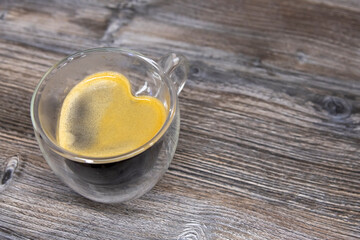 Double wall glass mug with fresh Caffè Americano stands on wooden table. Heart shaped coffee surface covered with crema. Copy space for your text. Romantic drink theme.