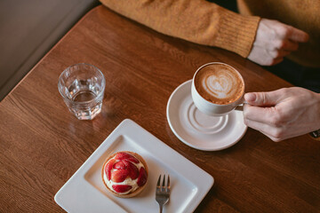 Cup of coffee, delicious cute strawberry tart on wooden table. Man having breakfast in cafe.