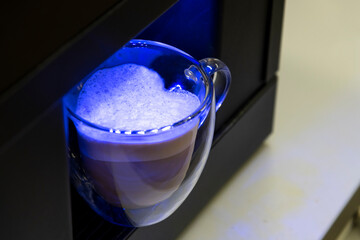 Double wall glass mug with fresh Caffè latte made from almond milk stands on black automatic coffee machine. Blue illumination. Heart shaped coffee surface. Selective focus. Hot drink theme.