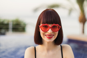 Portrait of young female with red hair wearing black swimsuit in swimming pool with red sunglasses enjoying sun and hot summer. Spa and sunbathing outdoor.