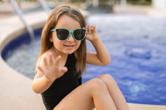 Little caucasian girl in sunglasses and black bathing suit waving hello, sitting by the pool in summer. Little cute girl relaxing in swimming pool. Summer travel hotel vacation - Image