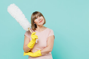 Elderly minded housewife woman 50s in pink t-shirt gloves doing housework hold white duster brush look aside isolated on plain pastel light blue background. Housekeeping cleaning tidying up concept.
