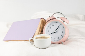Fototapeta na wymiar Pink alarm clock with cup of coffee and book on the bed close up image. Morning concept.