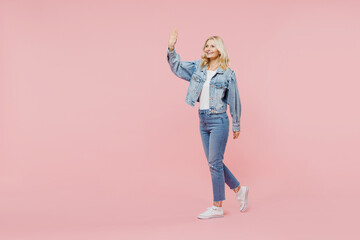 Full size side view elderly smiling fun happy woman 50s wearing denim jacket walking going waving hand say hello hi isolated on plain pastel light pink background. People lifestyle fashion concept