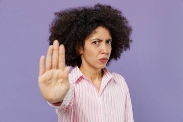 Young serious trict angry sad woman of African American ethnicity 20s wear pink striped shirt showing stop gesture with palm refusing isolated on plain pastel light purple background studio portrait