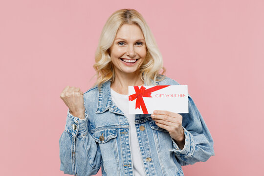 Elderly smiling happy woman 50s wear denim jacket hold card flyer voucher mock up for store do winner gesture isolated on plain pastel light pink background studio. People lifestyle fashion concept