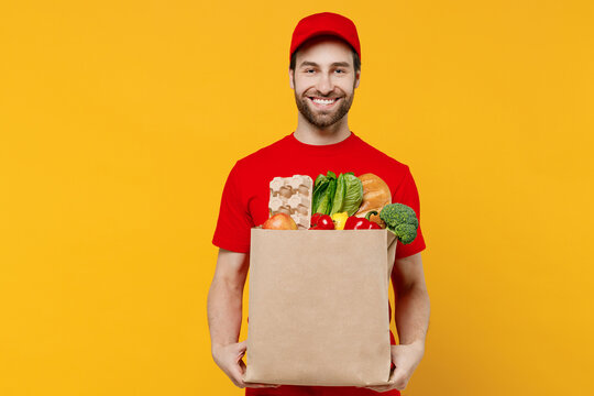 Smiling Delivery Guy Employee Man 20s Wear Red Cap T-shirt Uniform Workwear Work As Dealer Courier Hold Craft Brown Paper Bag With Grocery Food Vegetables Isolated On Plain Yellow Background Studio