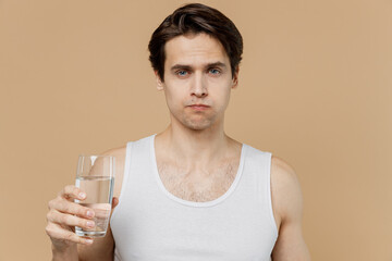 Attractive young man 20s perfect skin in undershirt hold glass of water drink rinses teeth isolated on pastel pastel beige background studio portrait. Skin care healthcare cosmetic procedures concept.