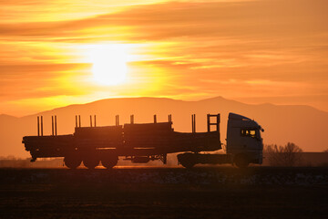 Fototapeta premium Semi-truck with cargo trailer driving on highway hauling cut down trees in evening. Timber delivery and logistics concept