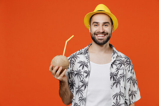 Young smiling happy tourist man wear beach shirt hat hold cocktail juice in coconut bowl with straw isolated on plain orange background studio portrait. Summer vacation sea rest sun tan concept.
