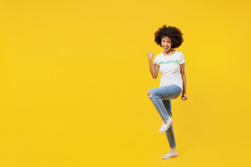 Fototapeta na wymiar Full body young woman of African American ethnicity wears white volunteer t-shirt do winner gesture raise up leg isolated on plain yellow background. Voluntary free work assistance help grace concept.